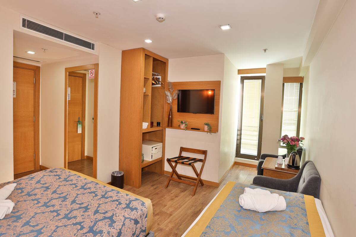 A hotel room with two beds, a TV mounted on the wall, a desk with a chair, and a small table with a vase of flowers.