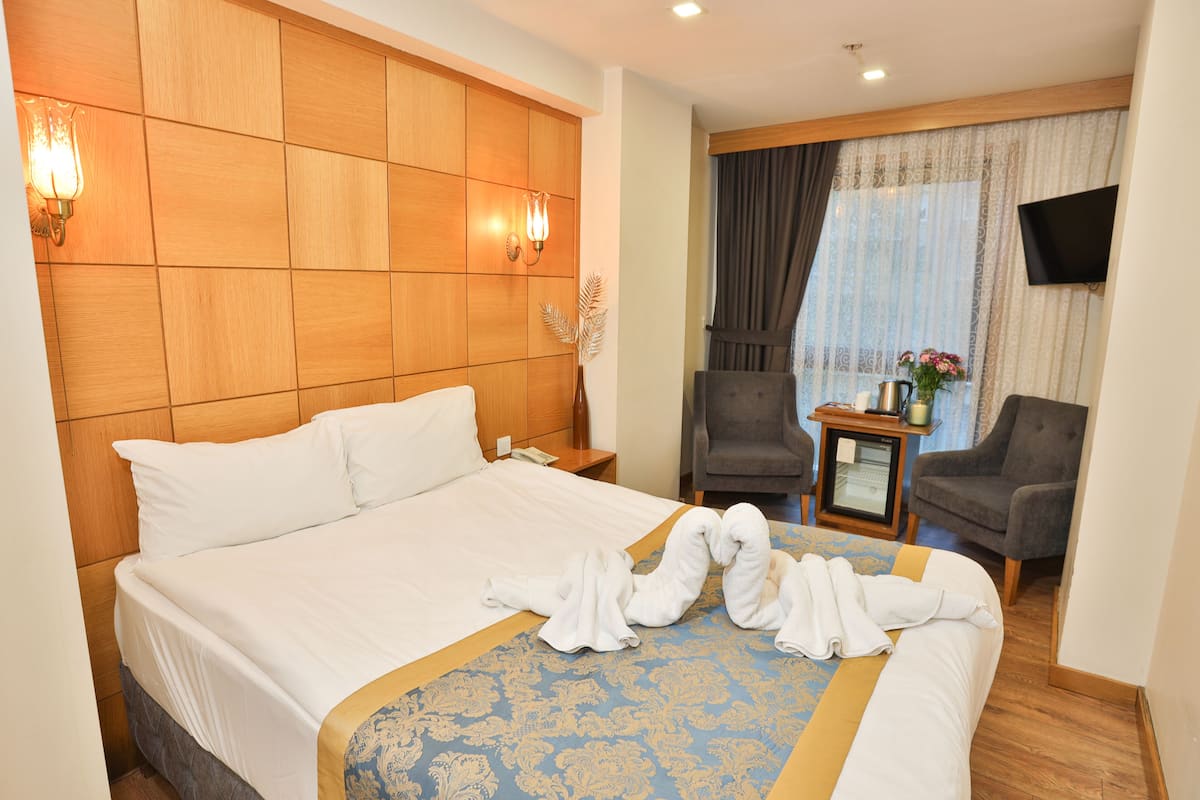A hotel room with a large bed, a wooden headboard, two wall-mounted lamps, a television, a small table with a pitcher and flowers, and a chair.