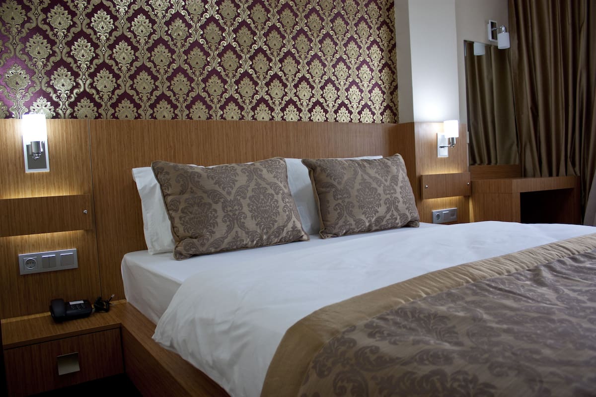 A hotel room with a wooden headboard, two patterned pillows, a white bedspread, bedside lamps, and a nightstand with a phone and charger.
