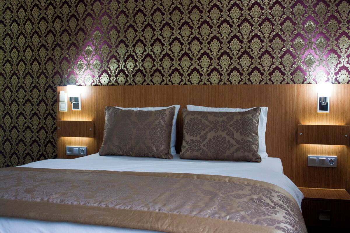 A hotel room with a large bed, two pillows, a wooden headboard, bedside lamps, and a patterned wallpaper.