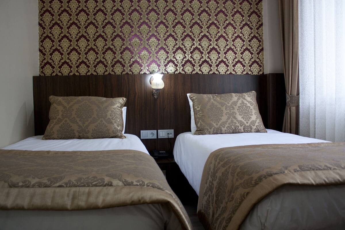 A hotel room with two beds, a patterned wall, a nightstand with a lamp, and a curtain.
