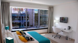Een hotelkamer met een groot raam, een bed met een blauwgroene deken, een bureau met een lamp en een stoel.