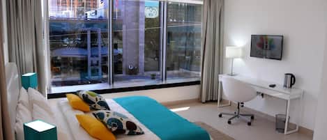 A hotel room with a large window, a bed with a teal blanket, a desk with a lamp, and a chair.