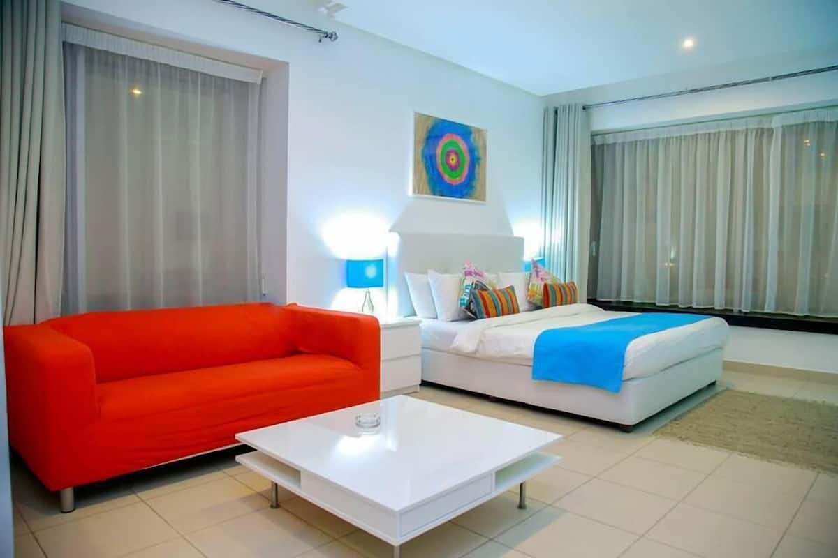 A modern hotel room with a red sofa, a white bed with colorful pillows, a white coffee table, and a target artwork on the wall.