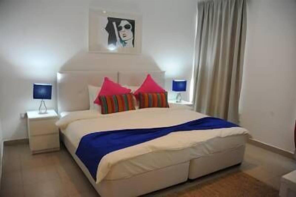 A neatly made bed with a blue blanket and multiple pillows, two bedside lamps, and a painting on the wall.