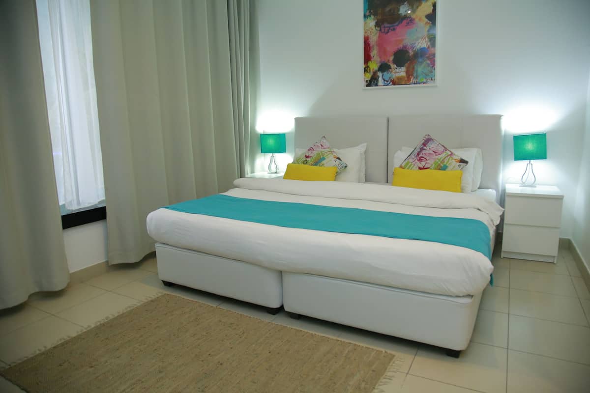 A bedroom with a large bed, two bedside lamps, a colorful painting on the wall, and a window with curtains.