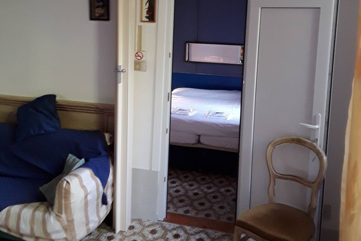 A room with a bed, a chair, a painting, a chandelier, and a door with a 'No Smoking' sign.