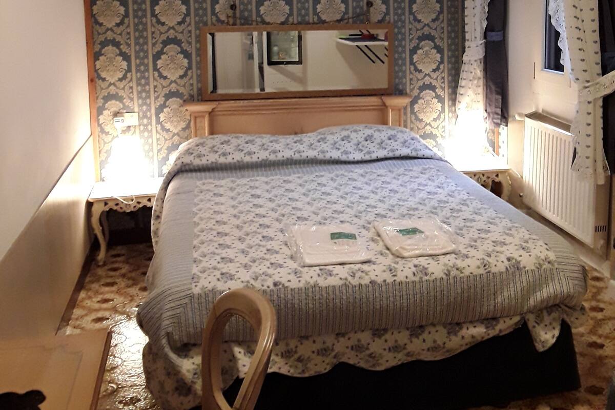 A bedroom with a bed, a wooden chair, a dresser, a mirror, and patterned wallpaper.