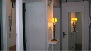 Een gang met een witte deur, een klein tafeltje met een lamp en een spiegel die de lamp weerspiegelt.