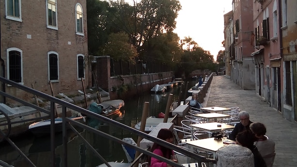 Een eetgedeelte aan het water met tafels en stoelen, een hekwerk en gebouwen met rondboogvensters.