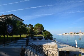 Un patio frente al mar con mesas al aire libre, un muro de piedra y vistas al océano y edificios a lo lejos.