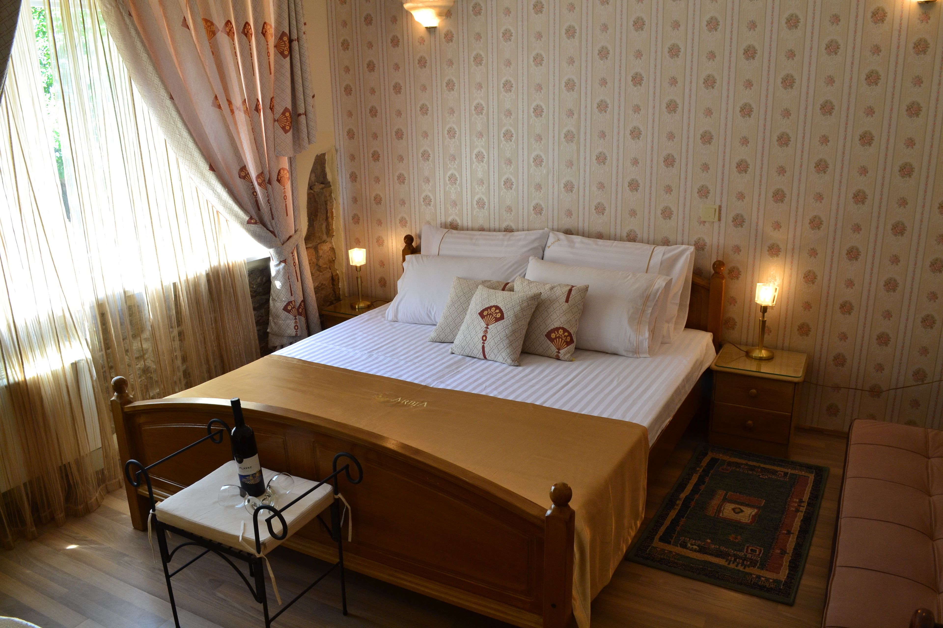 A neatly made bed with white linens and decorative pillows, a wooden nightstand with a lamp, a small table with a wine bottle, and a chair.