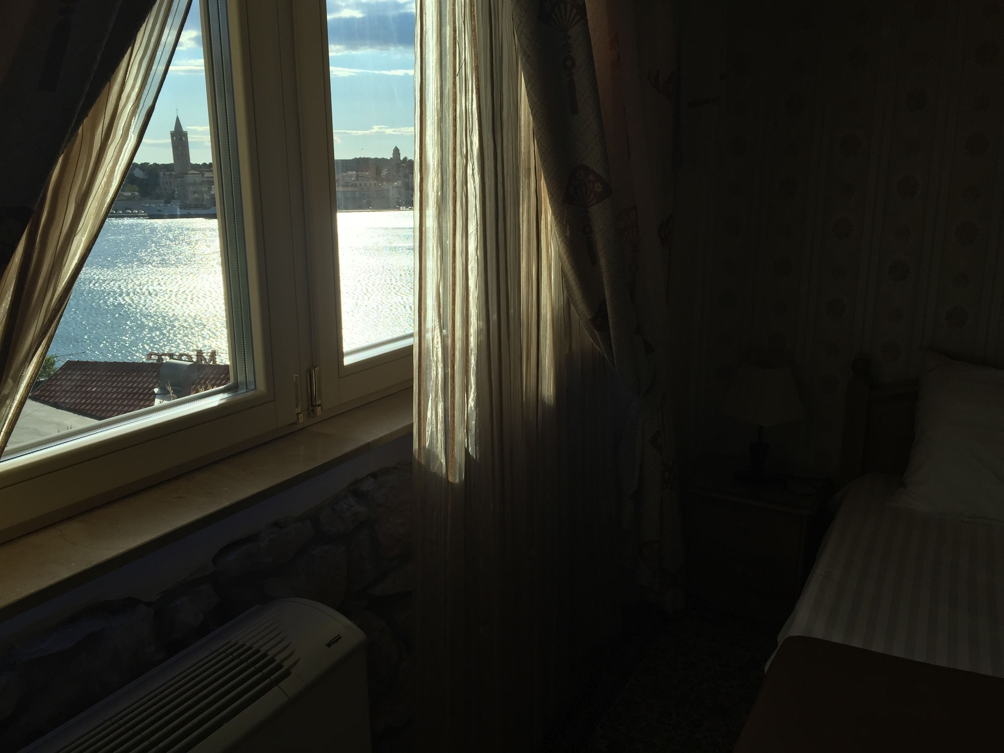 A room with a large window overlooking a body of water and a distant building. There is a bed with a striped bedspread and a radiator below the window.