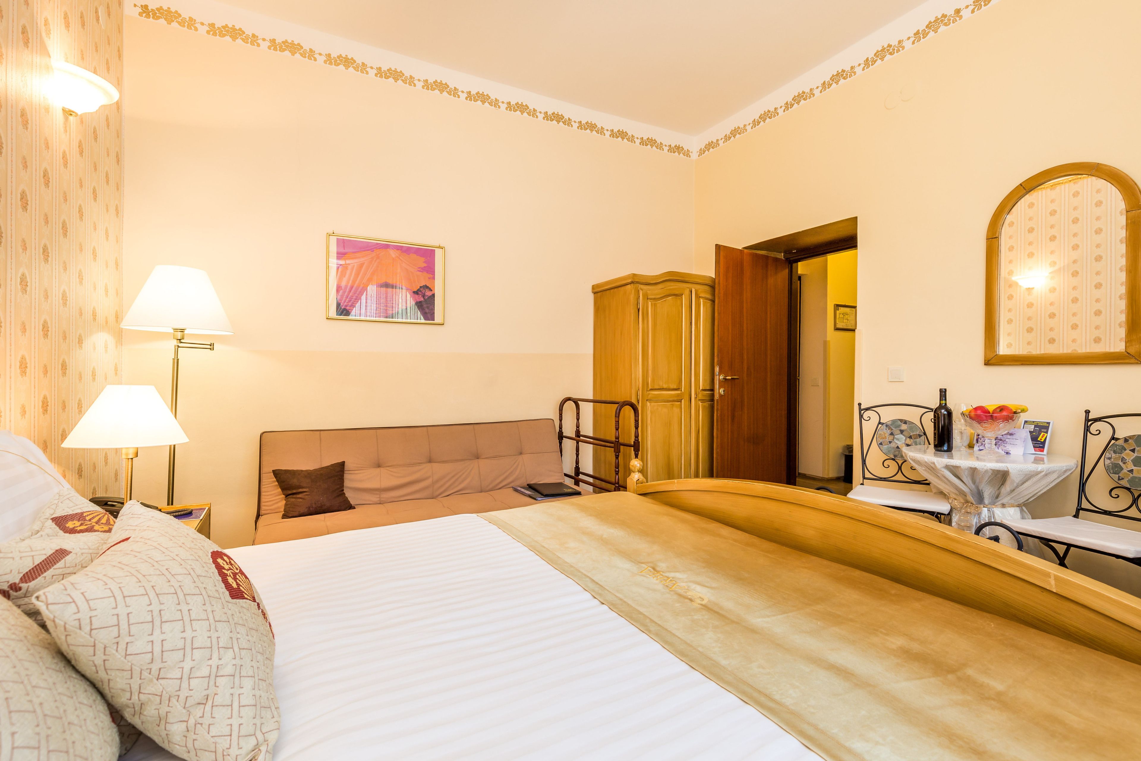 A hotel room with a large bed, a bedside table with a lamp, a wooden wardrobe, a small table with a vase of flowers, and a framed picture on the wall.