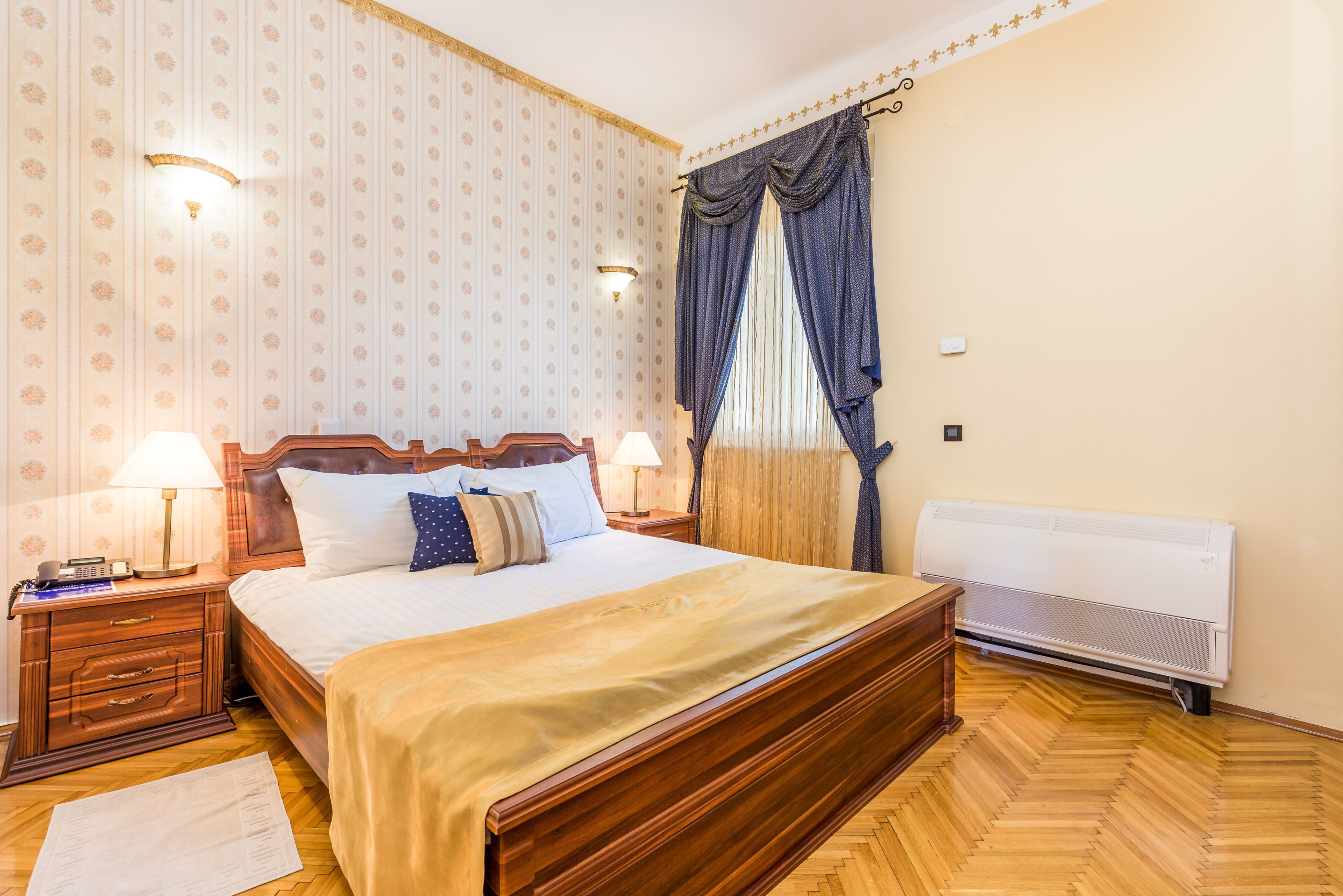 Una habitación de hotel con una cama de madera, dos mesitas de noche con lámparas, un radiador y un papel tapiz estampado.