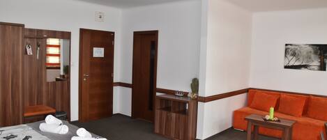 A hotel room with a bed, a wooden wardrobe, a wooden desk, a small table, an orange sofa, and a painting on the wall.