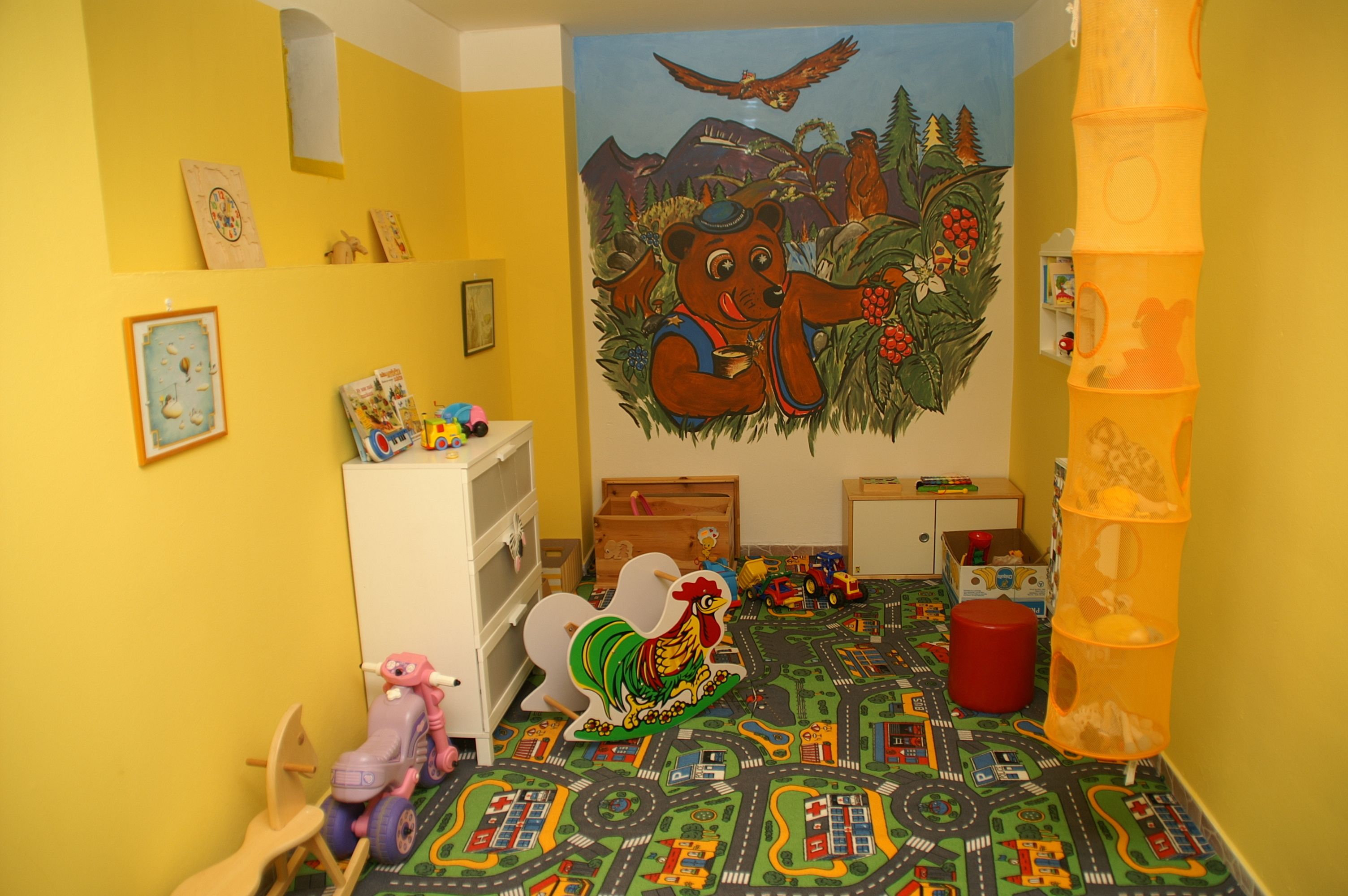 Un cuarto de juegos infantil con una alfombra de diseño vial colorido, un mural grande de un oso y varios juguetes.