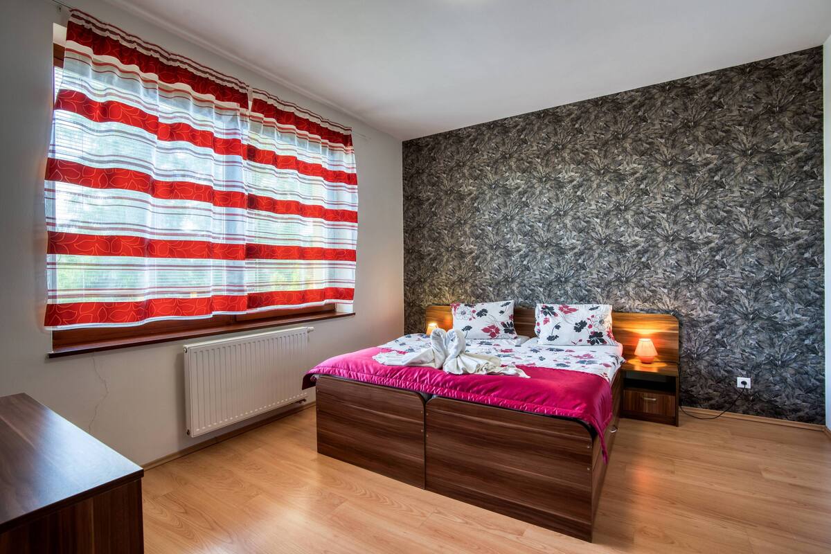 A bedroom with a wooden bed, a radiator, a window with red curtains, a nightstand with a lamp, and a textured wall.