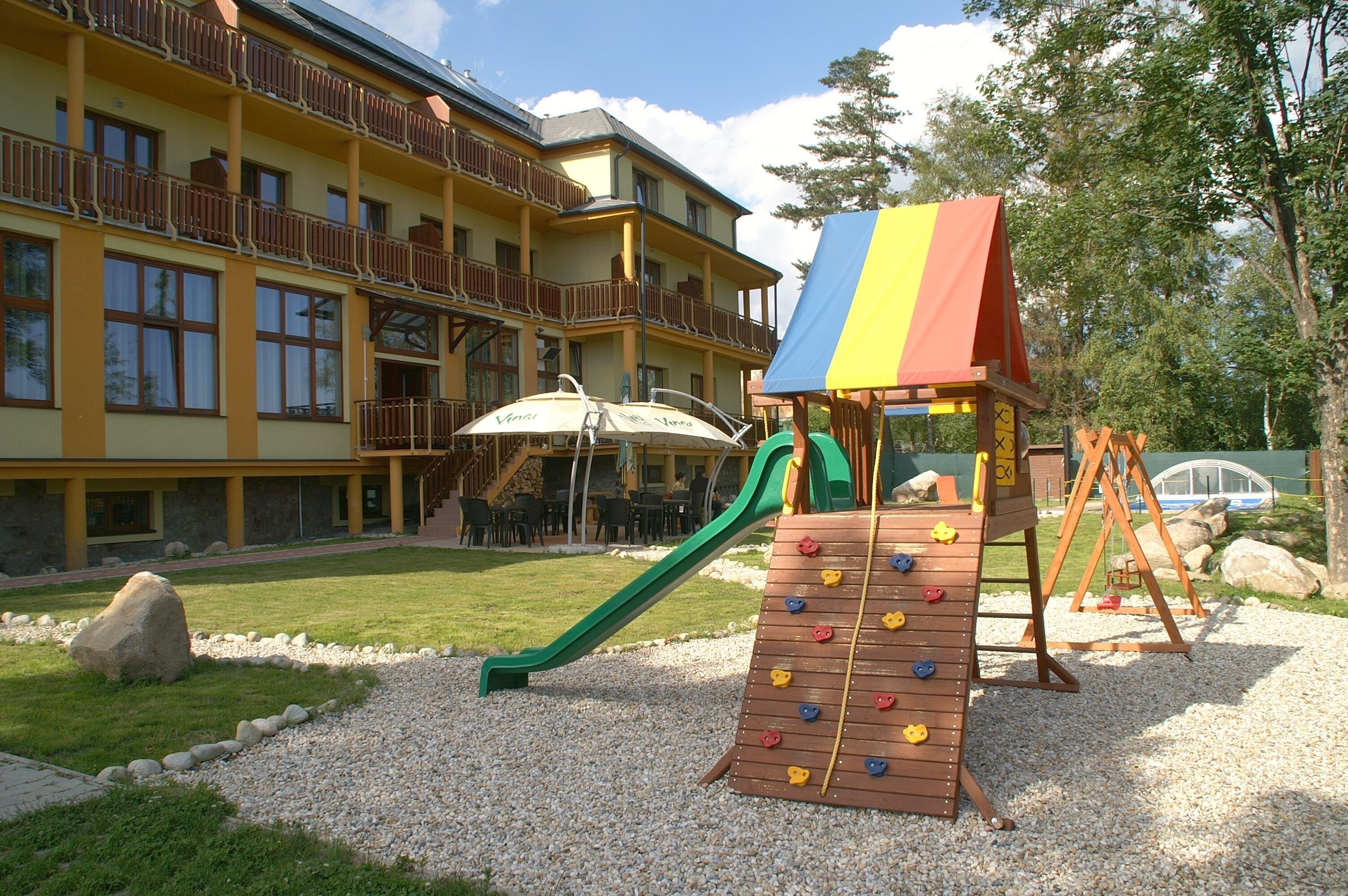 A playground with a climbing wall and slide, surrounded by a building with balconies and outdoor seating.