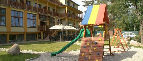 A playground with a climbing wall and slide, surrounded by a building with balconies and outdoor seating.