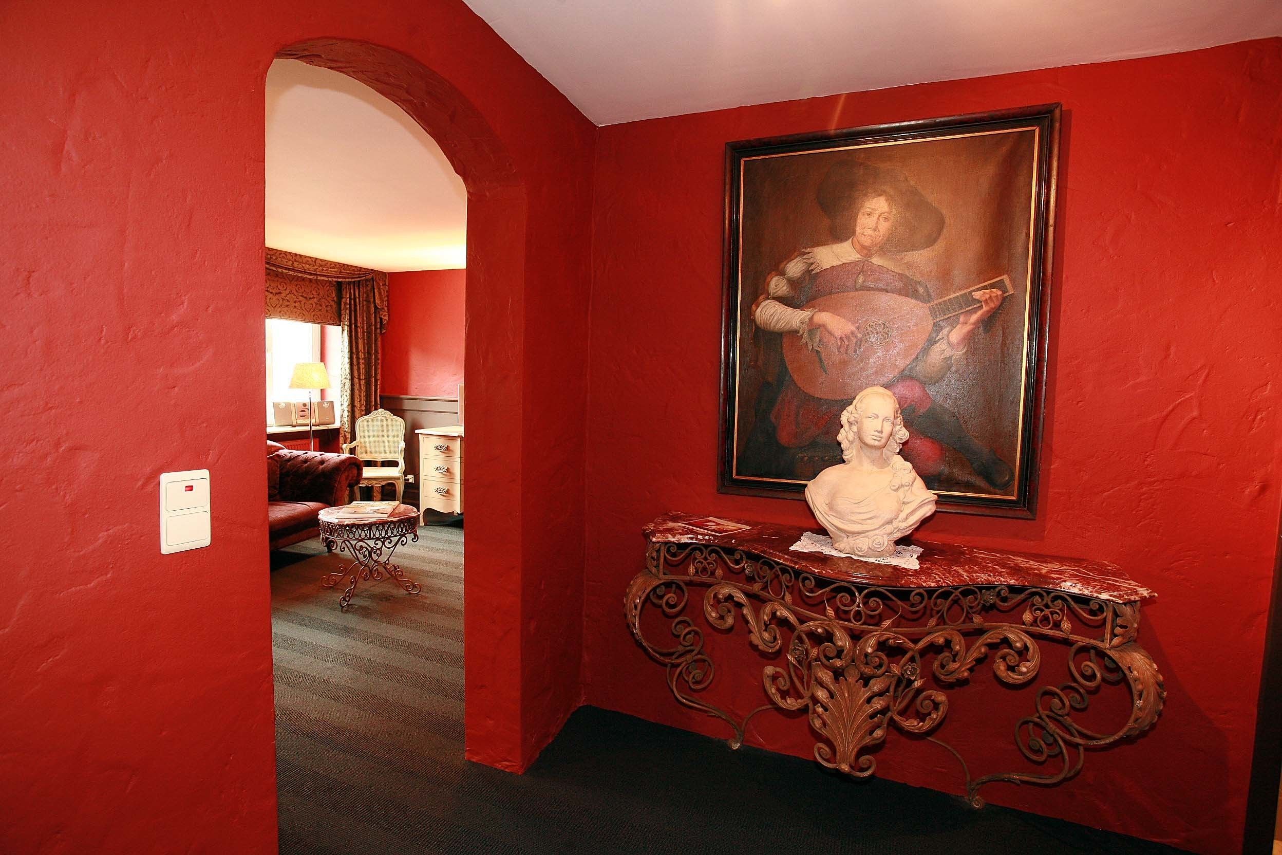 Une pièce aux murs rouges, ornée d’un tableau encadré, d’un buste blanc posé sur une table richement décorée, et d’une porte-fenêtre donnant sur une autre pièce.