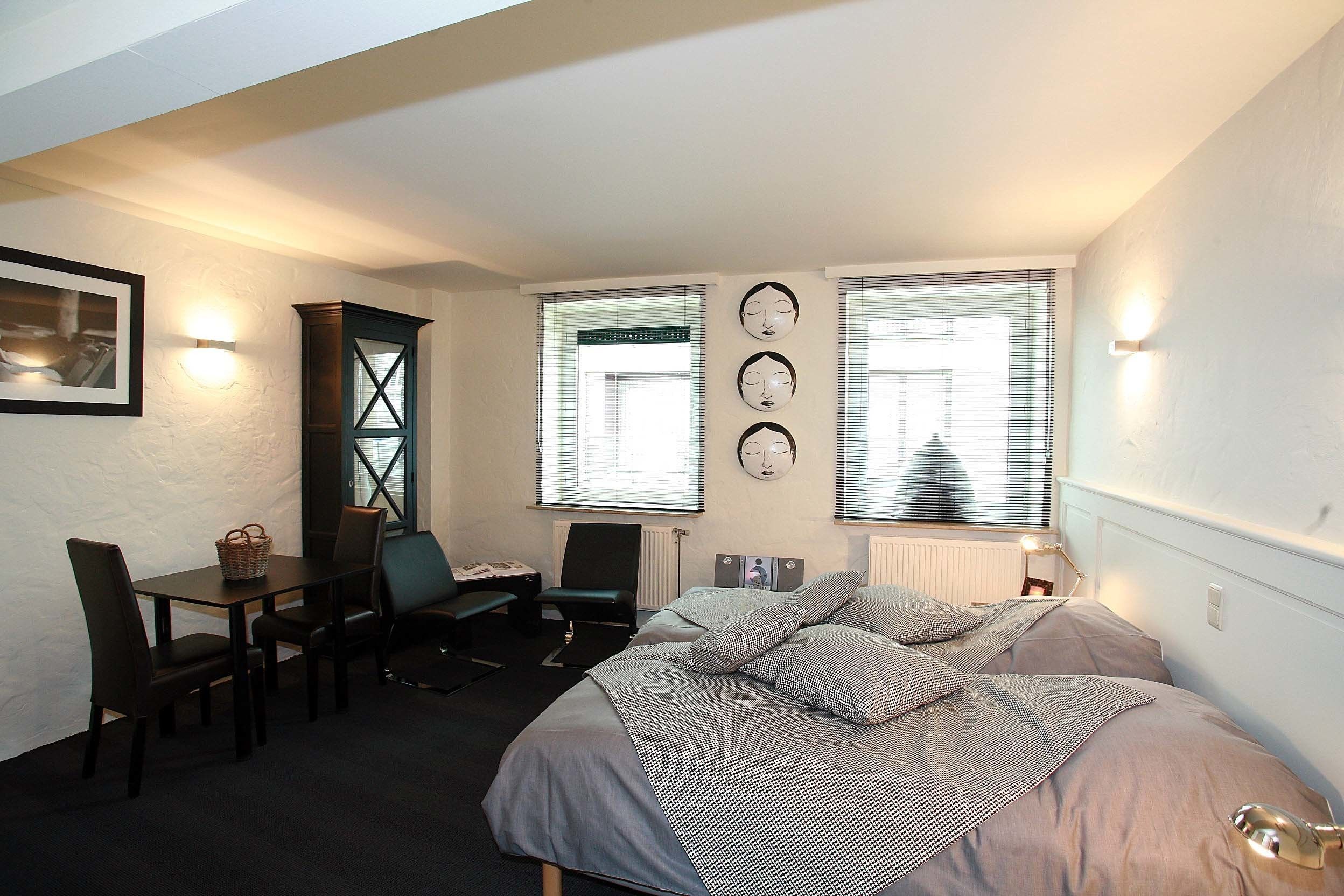 Une chambre à coucher avec un lit, un bureau et une chaise. Il y a deux fenêtres avec des stores, un radiateur et des lampes fixées au mur.