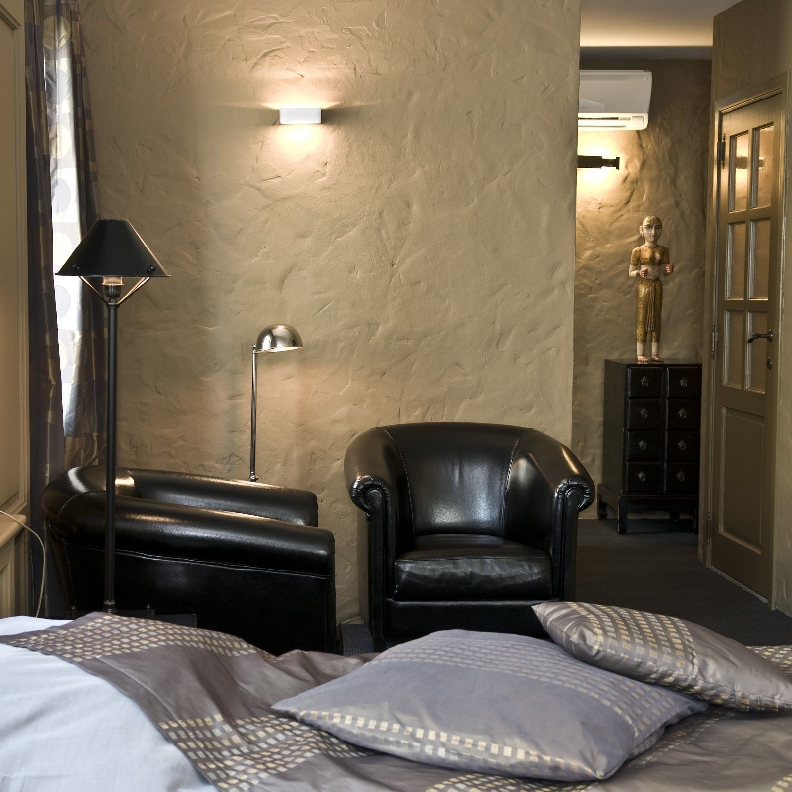 A hotel room with a bed, a black leather armchair, a floor lamp, a statue, and a dresser.