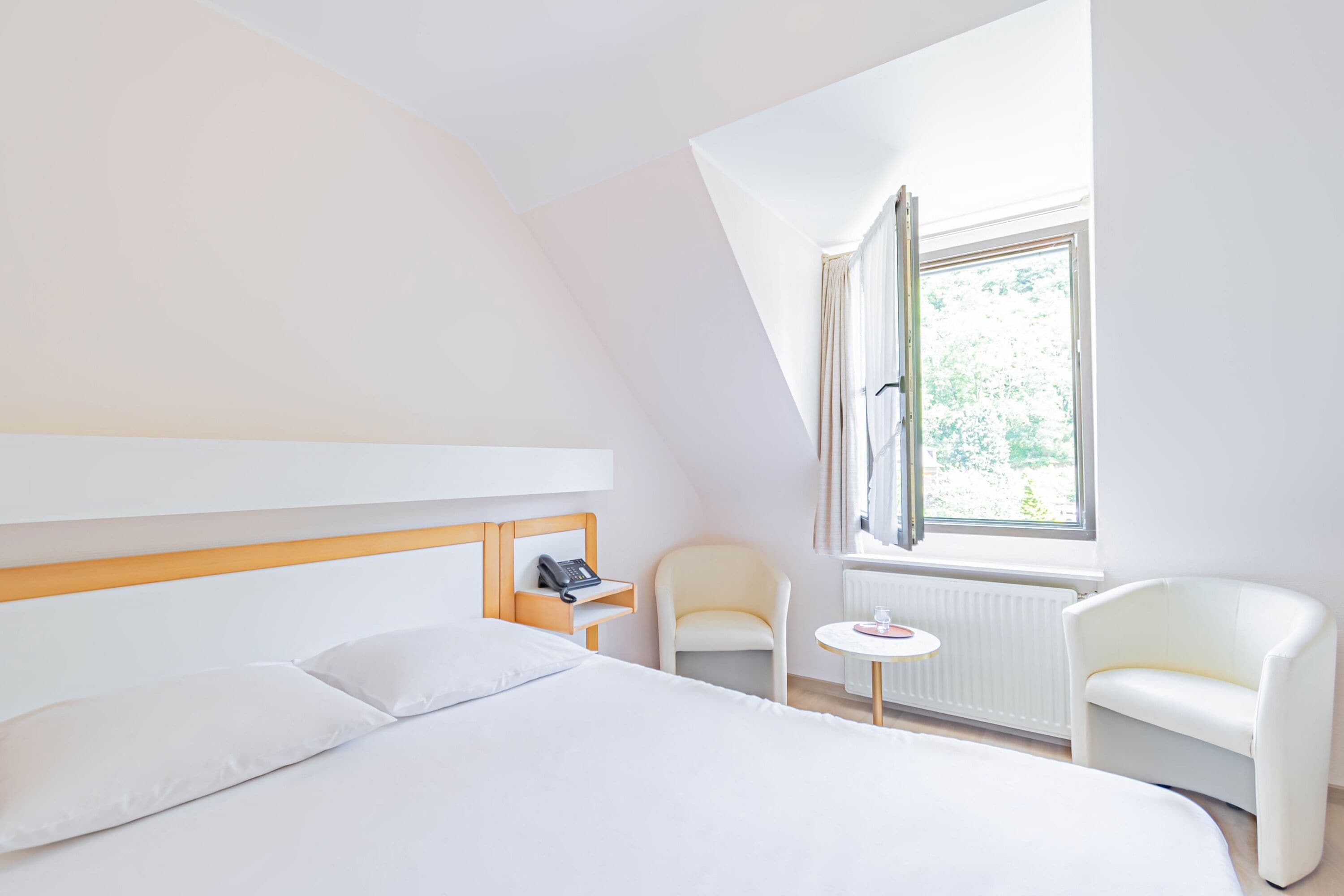 A hotel room with a bed, a chair, a small table, and a window with curtains.