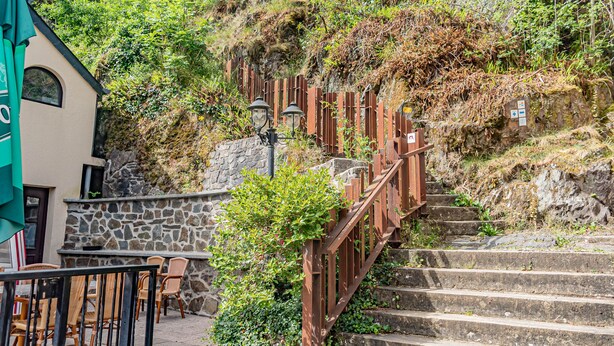 Una escalera de piedra que sube una colina, con barandilla de madera y un edificio con muro de piedra y una ventana en la parte superior.
