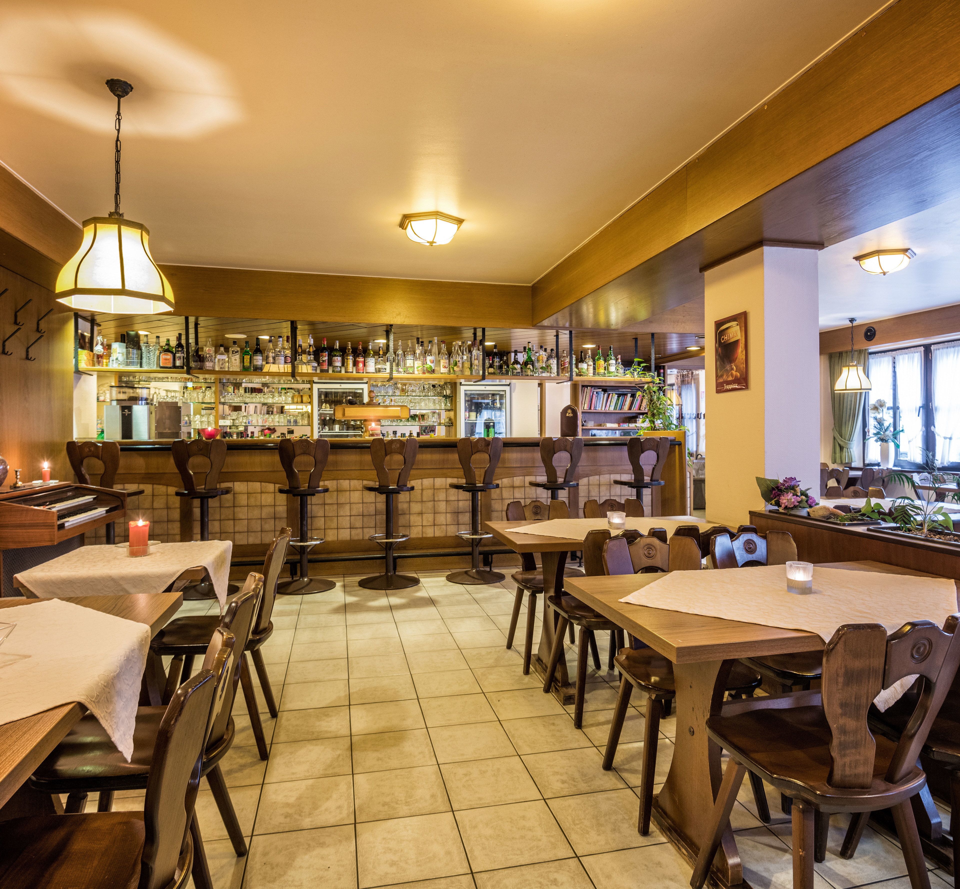 A restaurant interior with multiple tables and chairs, a bar area with shelves of bottles, and a tiled floor.