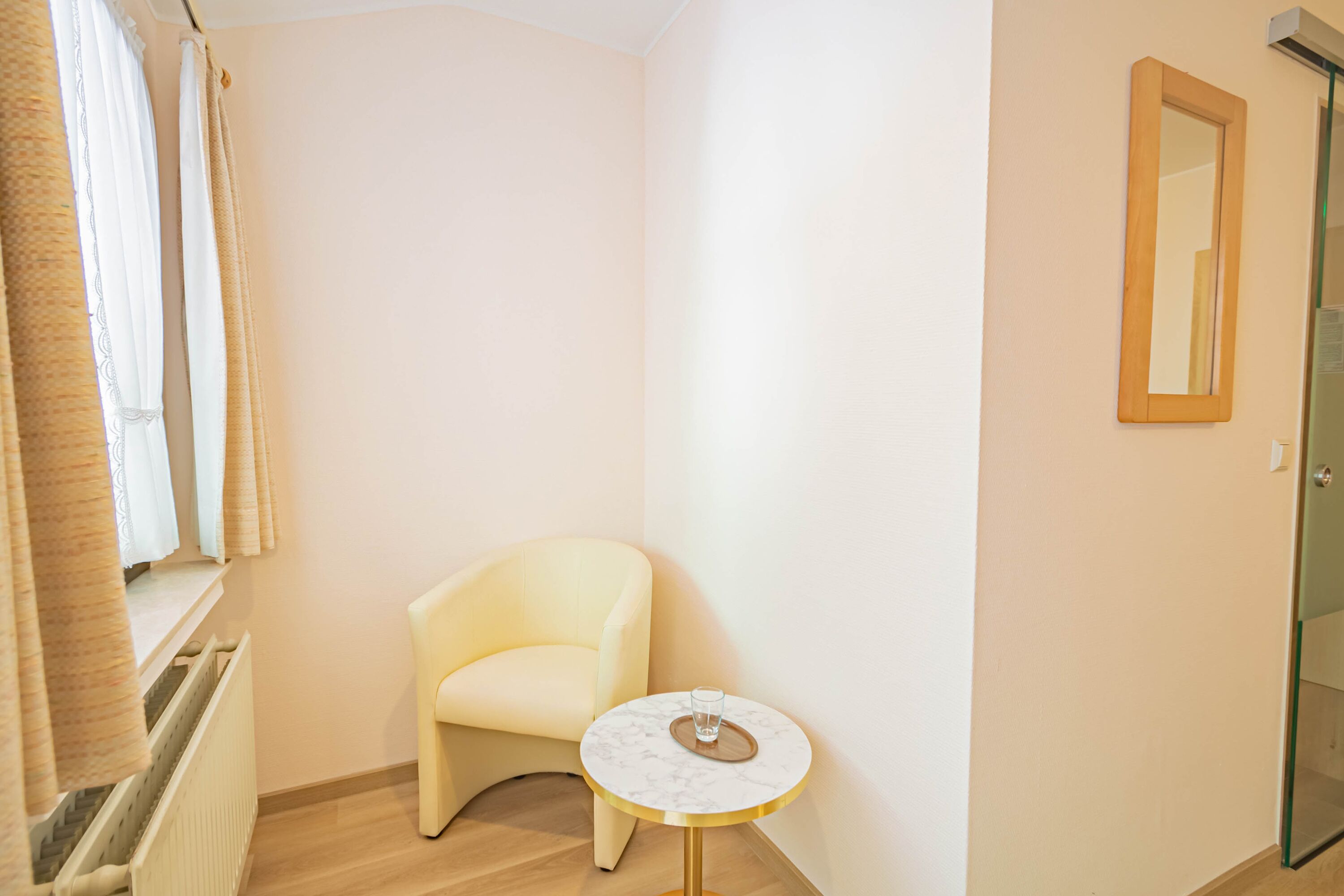 Een hoekappartement met een witte fauteuil, een klein rond tafeltje met een glas, en een spiegel aan de muur.