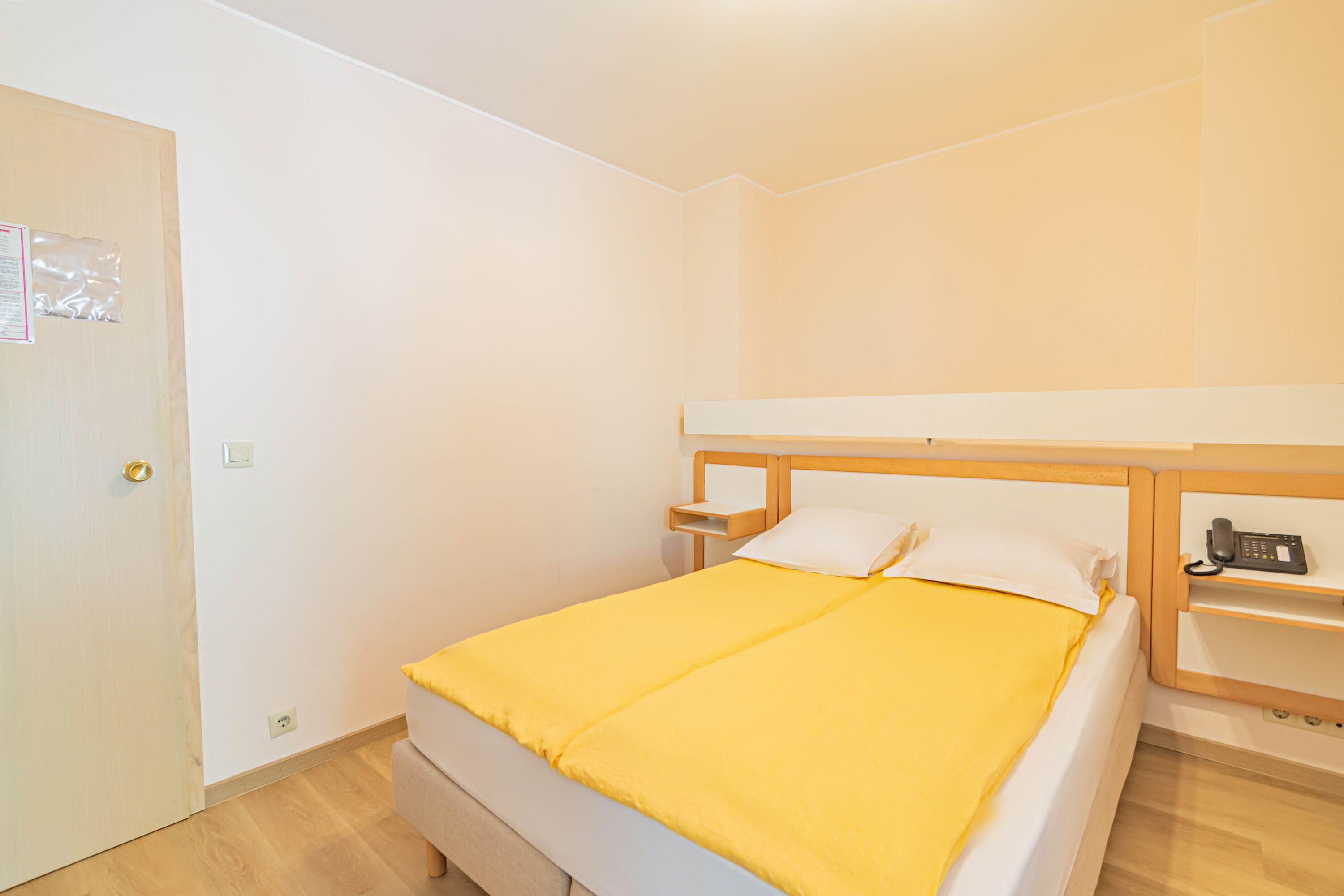 Habitación individual con una cama de una sola persona, cabecera de madera, mesita de noche con teléfono y colcha amarilla.