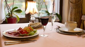 Una mesa dispuesta con un plato de comida, dos copas de vino y una taza, con una lámpara y plantas en macetas al fondo.