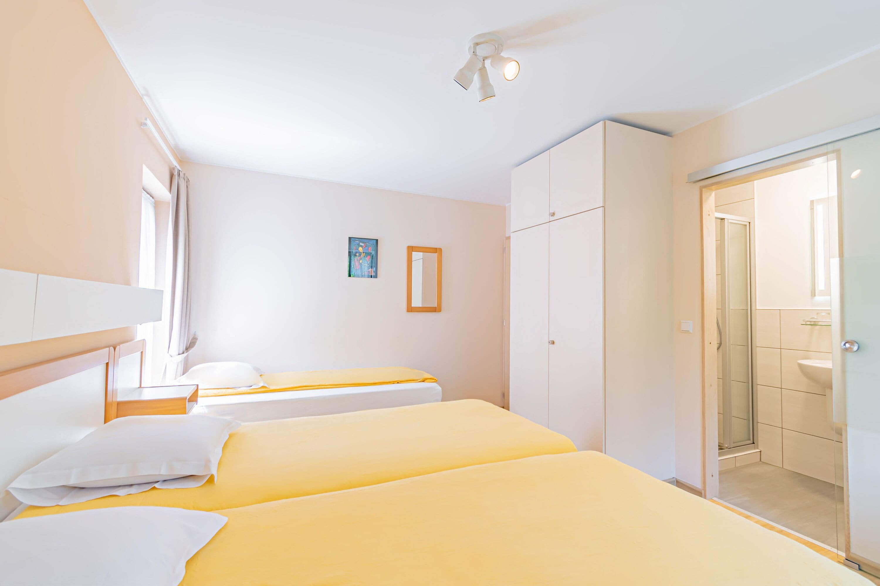 A hotel room with two beds, a wardrobe, a bathroom with a glass shower, and a painting on the wall.