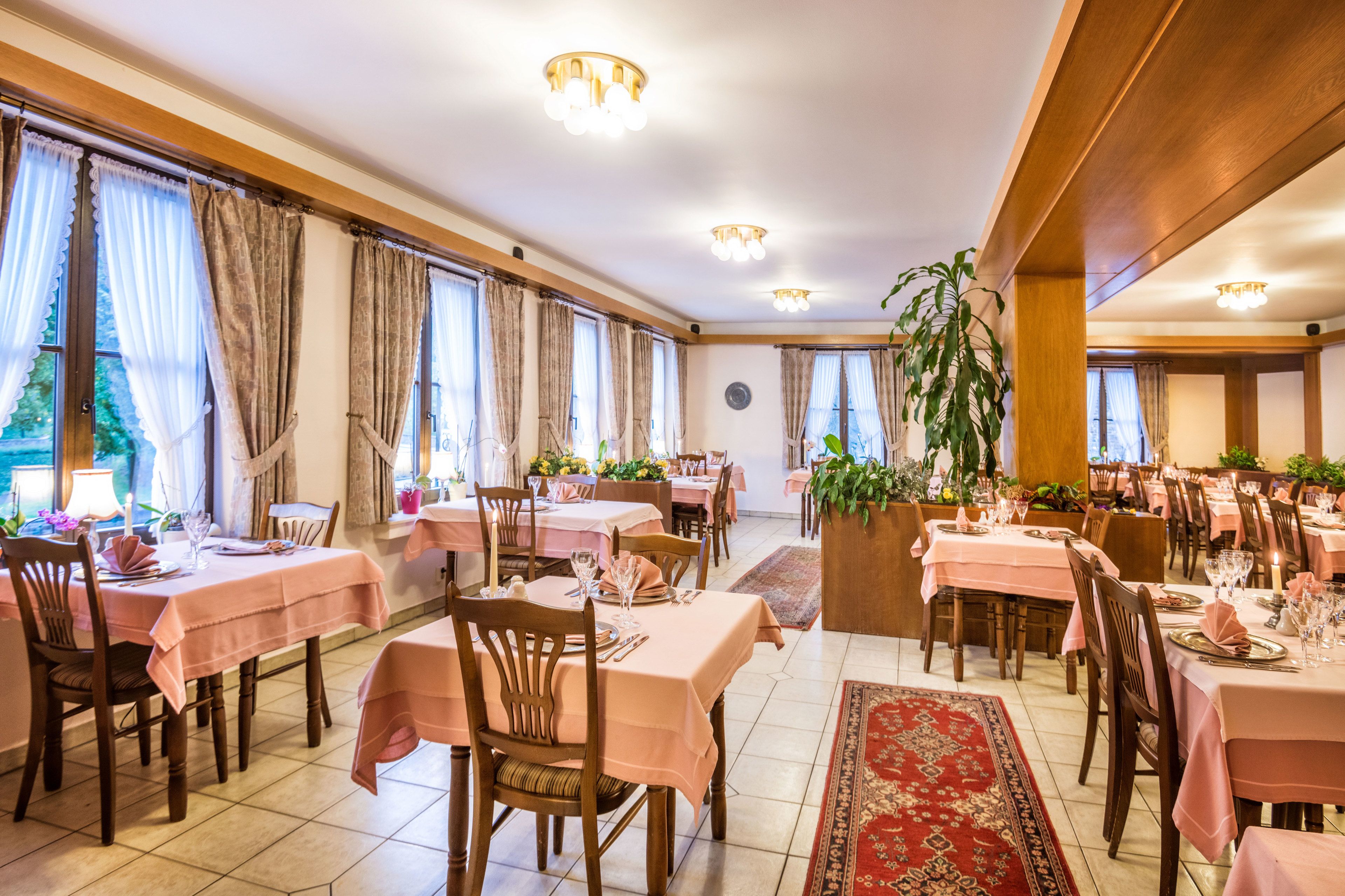 Un comedor con mesas dispuestas para el servicio, amplias ventanas con cortinas y una planta en el centro.