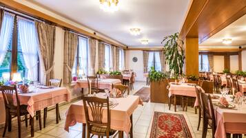 Un comedor con mesas dispuestas para el servicio, amplias ventanas con cortinas y una planta en el centro.