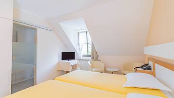 Een moderne hotelkamer met een schuin dak, twee bedden met geel-witte beddengoed, een bureau met een computer, een stoel en een klein tafeltje.