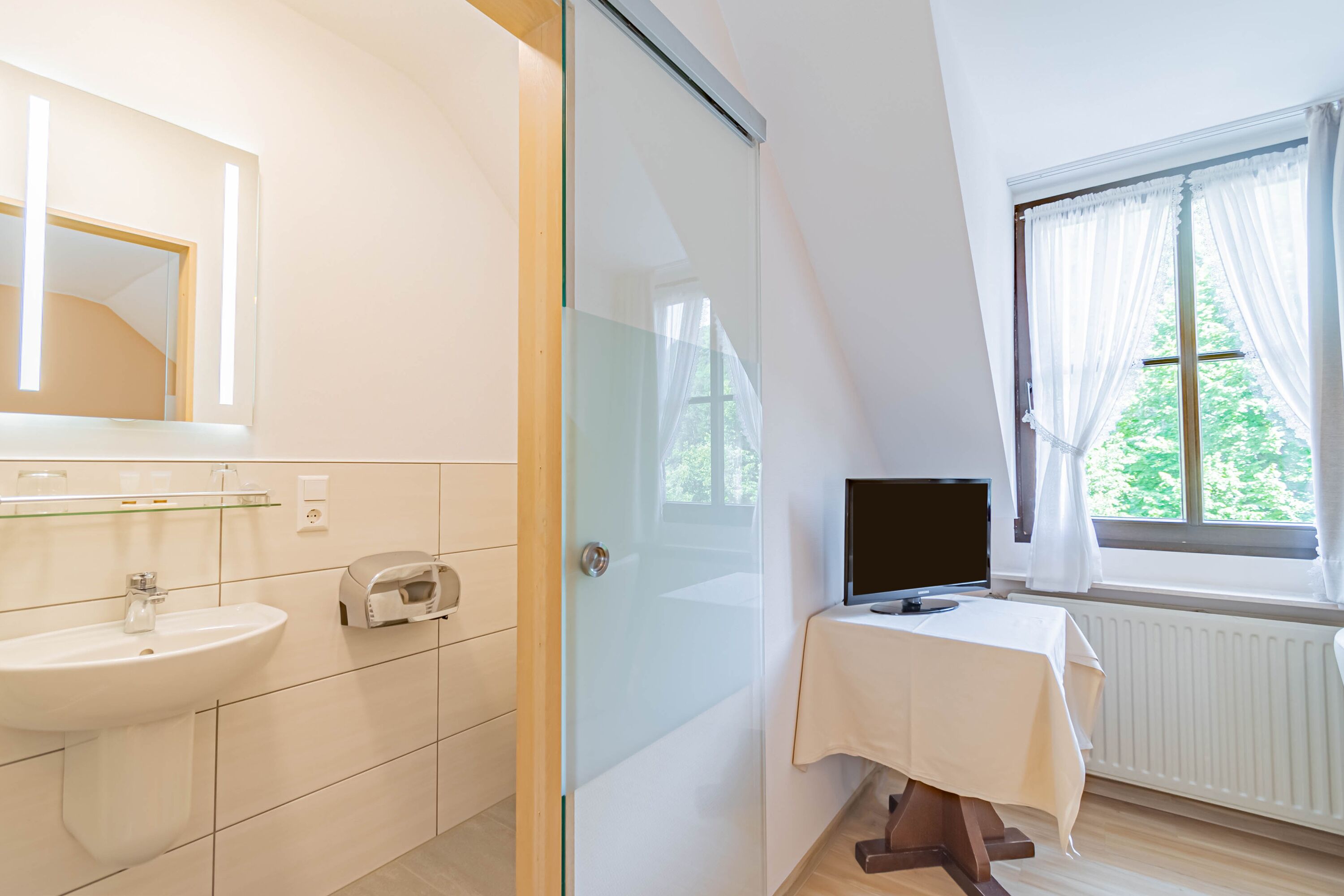 A modern bathroom with a glass shower enclosure, a sink, and a toilet. There is a small table with a TV and a window with curtains.