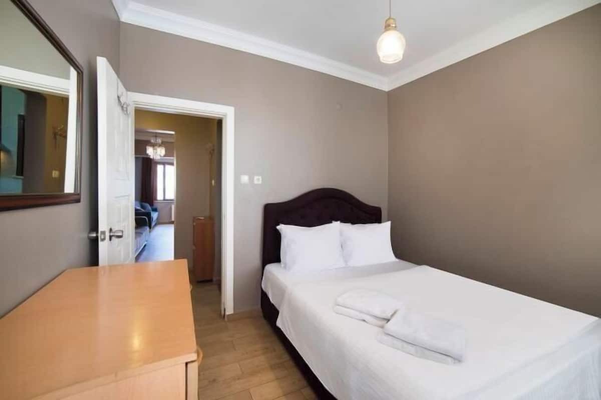 A bedroom with a bed, a wooden dresser, a mirror, and a door leading to another room.