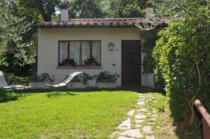 Una casita con techo de tejas, una puerta y ventanas con cortinas. Hay un sendero que lleva a la casa y una silla en el patio.