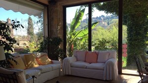 A cozy living room with a white sofa, a cushioned chair, and a large window offering a view of greenery and a distant building. - Relais San Bruno (Montepulciano)