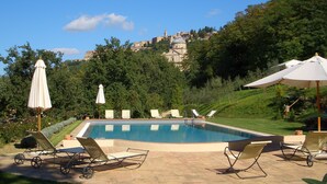 A pool area with lounge chairs and umbrellas, surrounded by greenery and a view of a hill with buildings. - Relais San Bruno (Montepulciano)