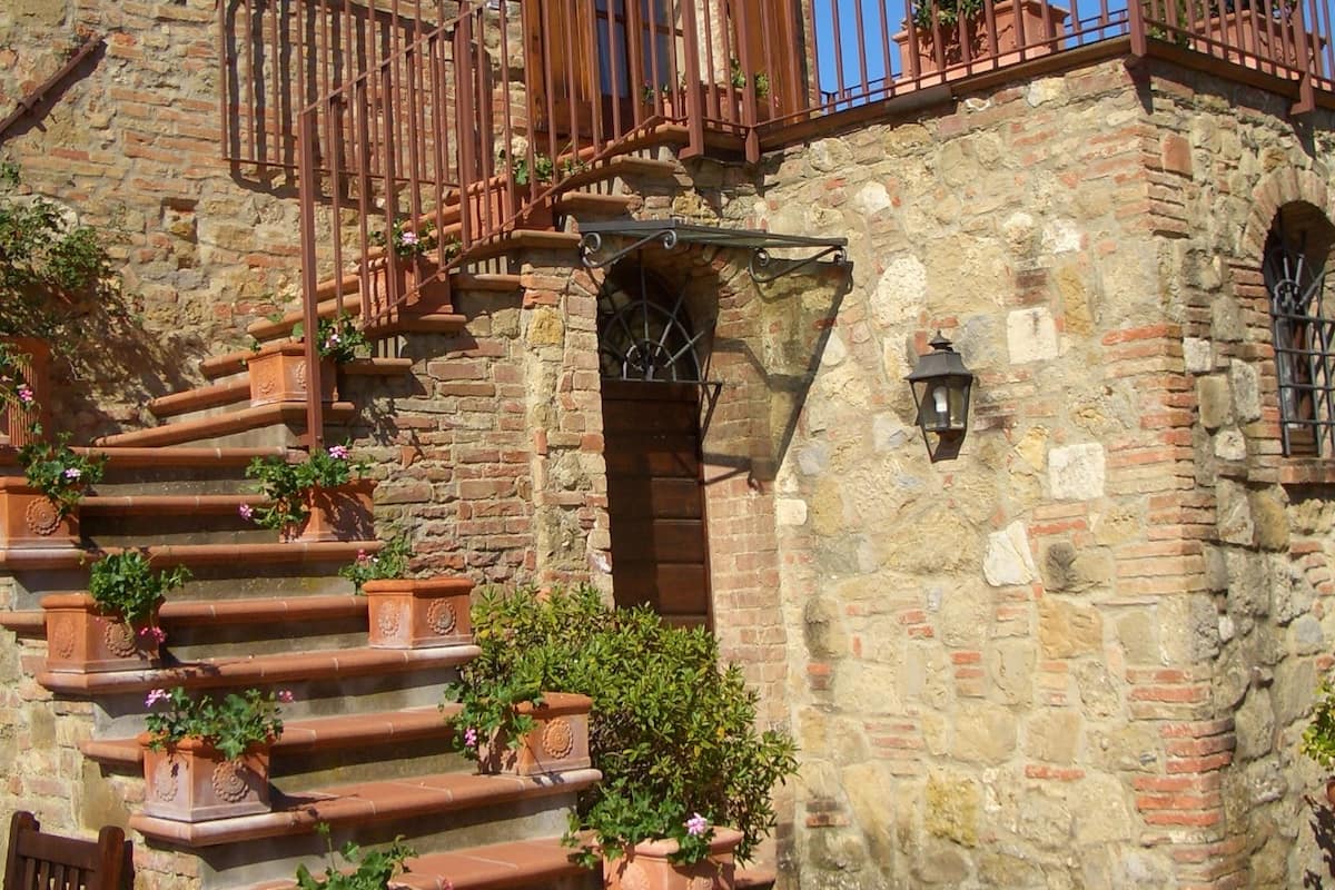 Ein Steinhaus mit einer Treppe, die zu einem hölzernen Balkon führt, umgeben von Pflanzkübeln.