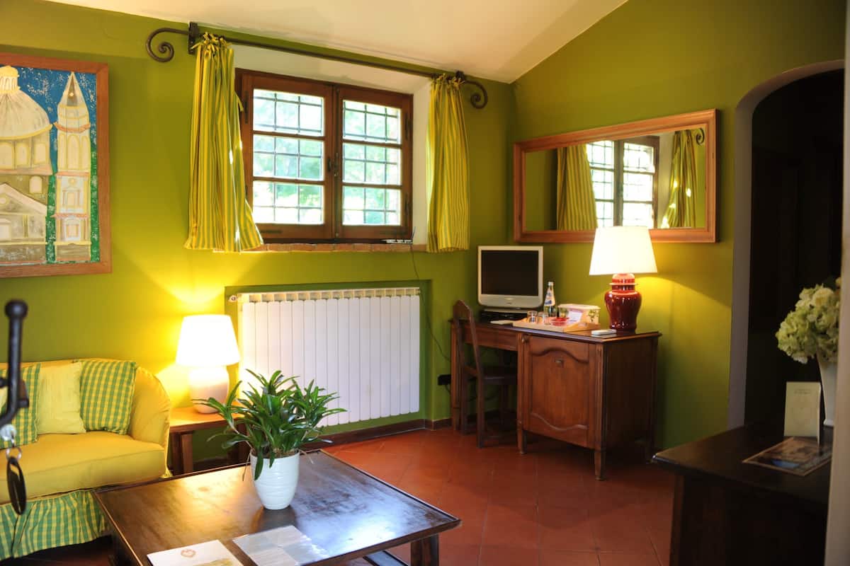 Ein Zimmer mit grüner Wand, einem Fenster mit Vorhängen, einem Schreibtisch mit Computer, einer Lampe, einer Pflanze und einem Sofa.