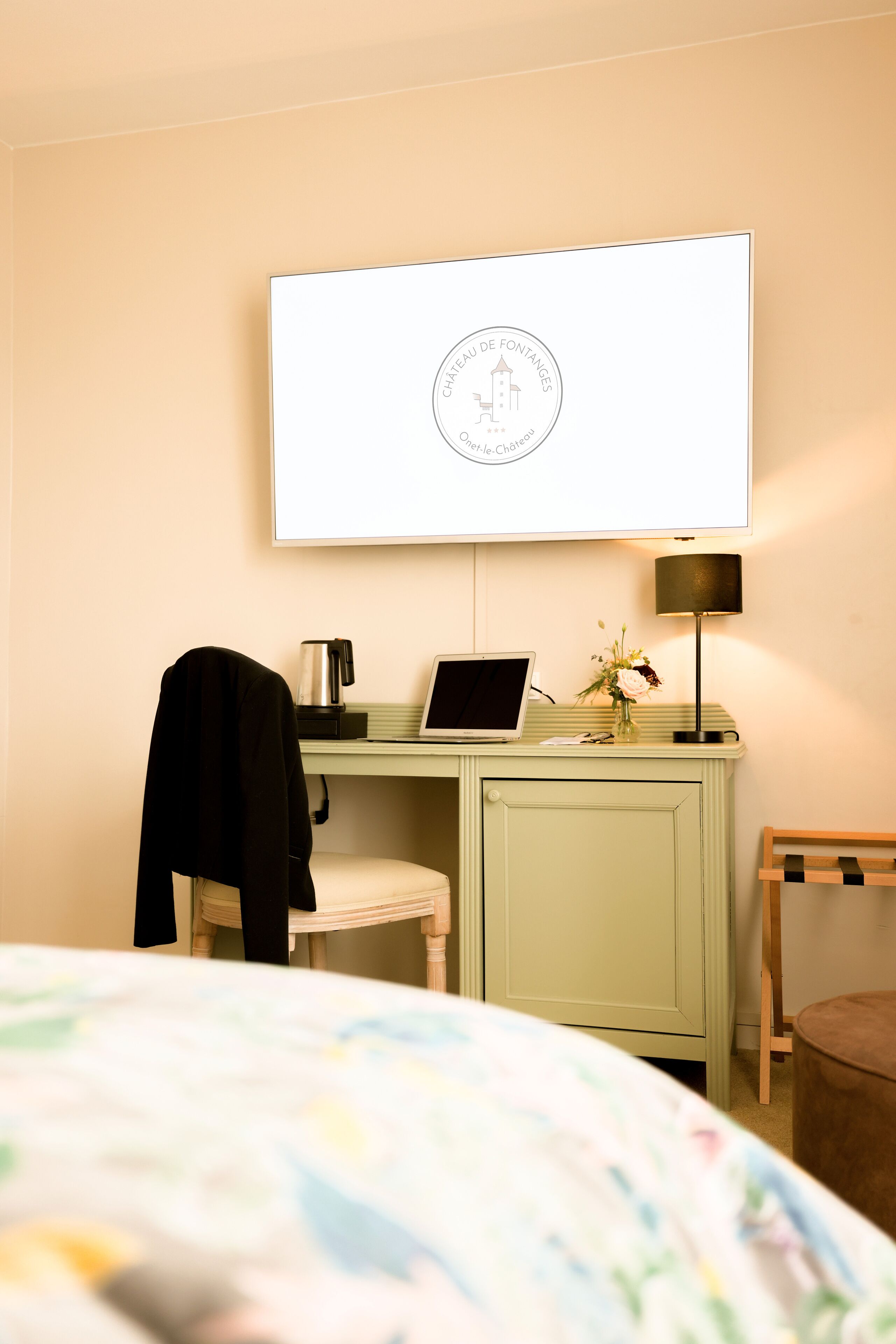 A hotel room with a bed, a desk with a laptop, a coffee maker, a lamp, and a TV displaying a logo.