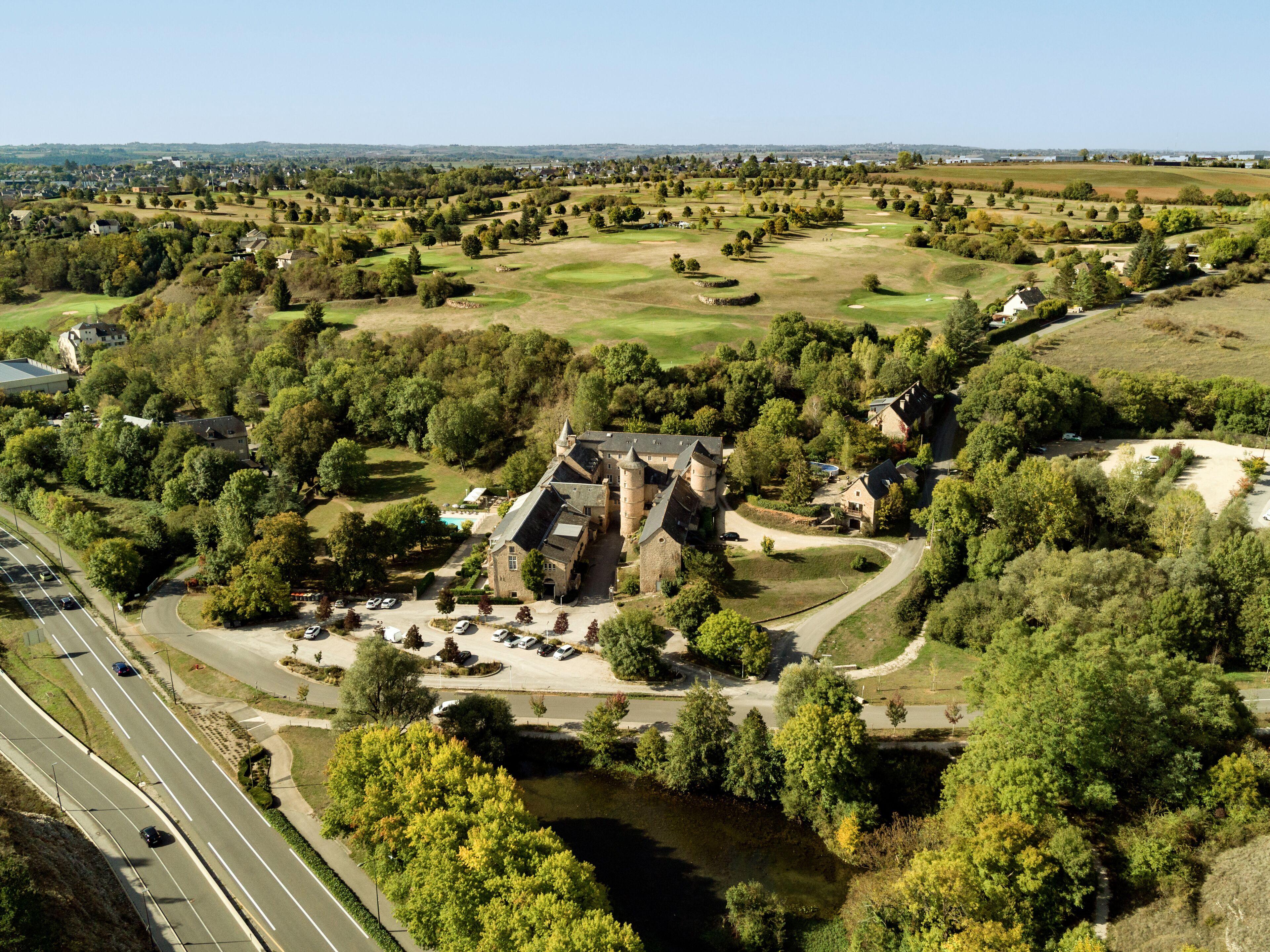 En slotlignende bygning omgivet af frodig vegetation, en parkeringsplads og en golfbane.