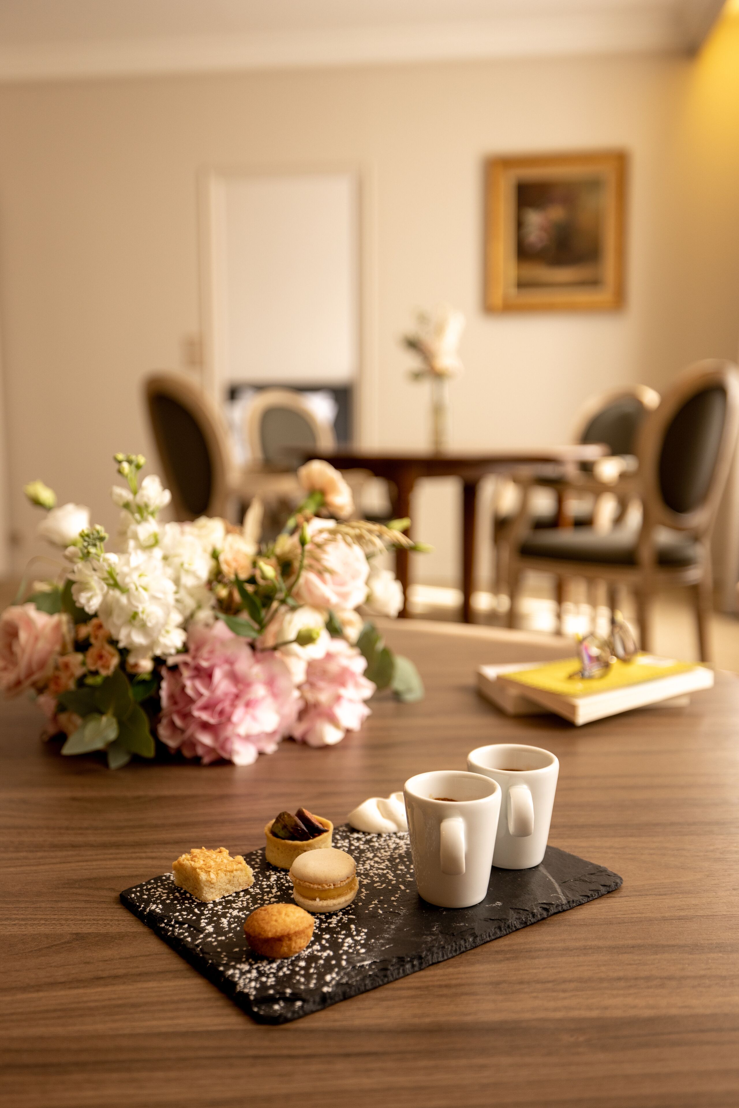 Una mesa de comedor con un arreglo floral, macarons y dos tazas de café sobre un posavasos negro.