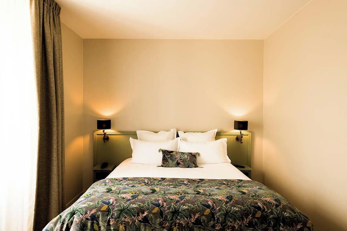 A hotel room with a bed, two bedside lamps, a floral bedspread, and a curtain.