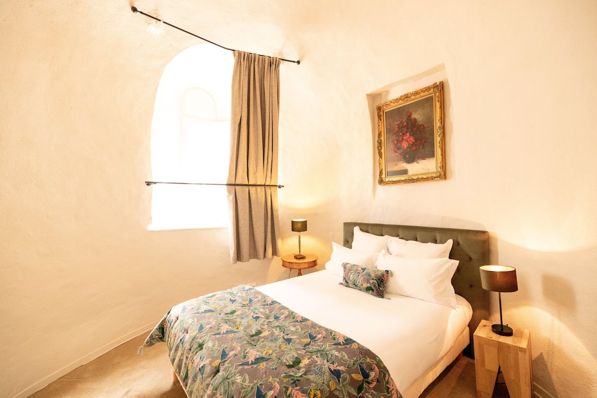 A bedroom with a bed, a nightstand, a lamp, a window with curtains, and a framed picture on the wall.