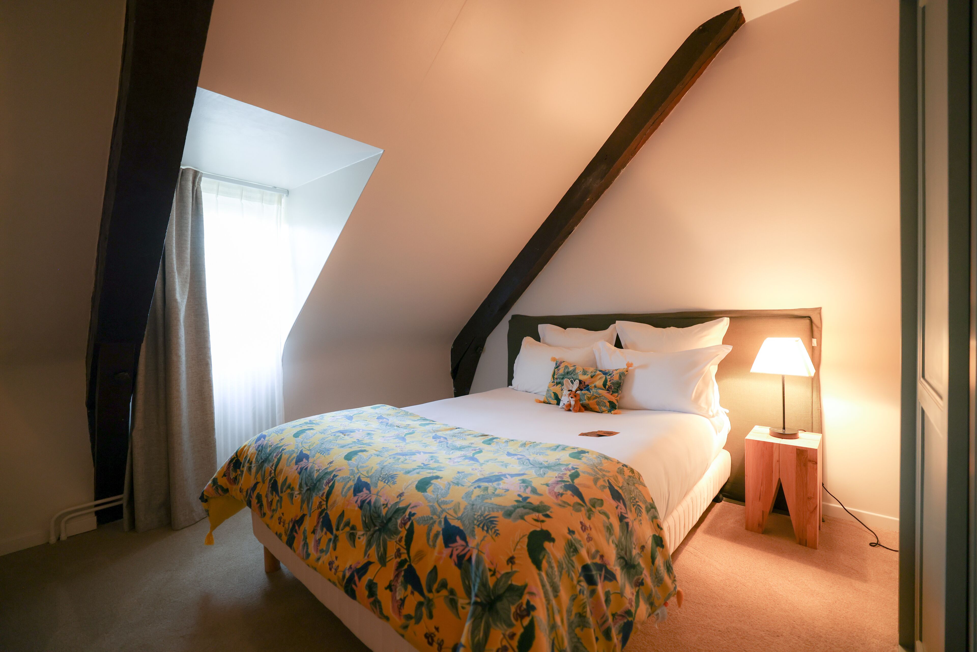 A bedroom with a sloped ceiling, a bed with a floral bedspread, white pillows, a nightstand with a lamp, and a window with curtains.